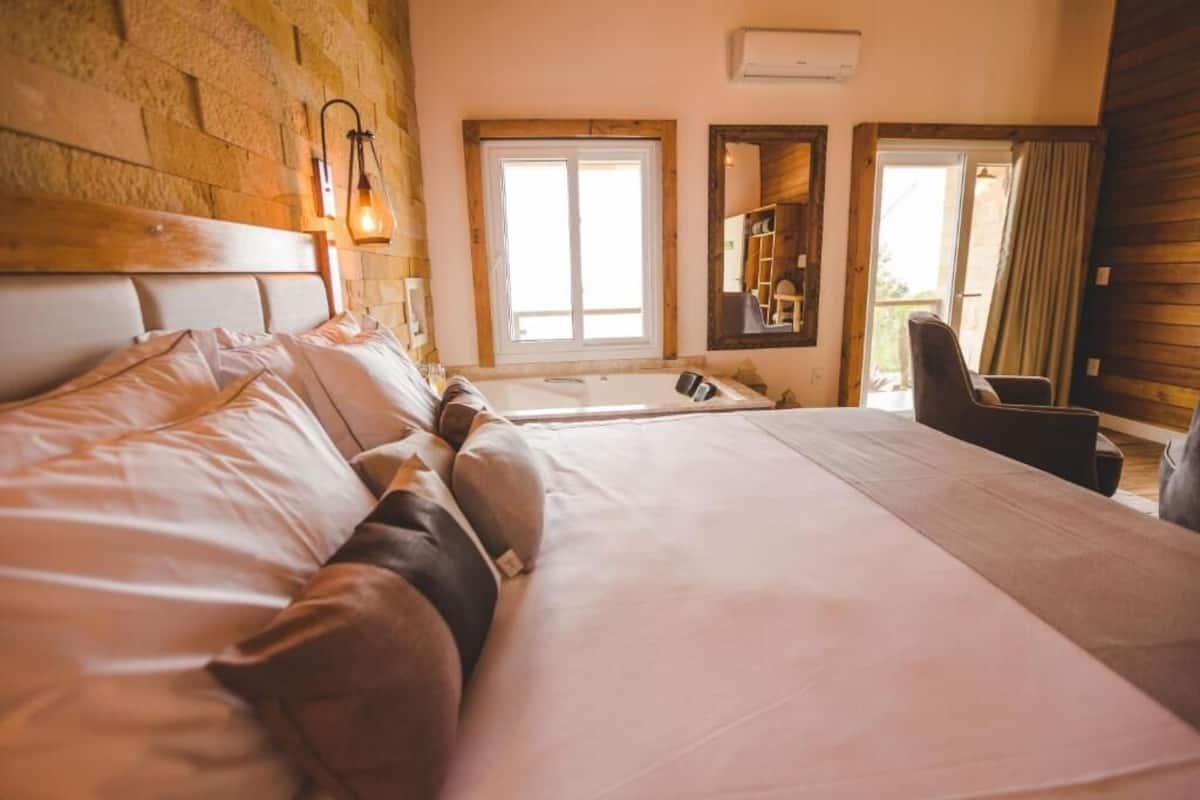 A bedroom with a large bed, a wooden headboard, a small table, and a chair. There are windows with curtains and a mirror on the wall.