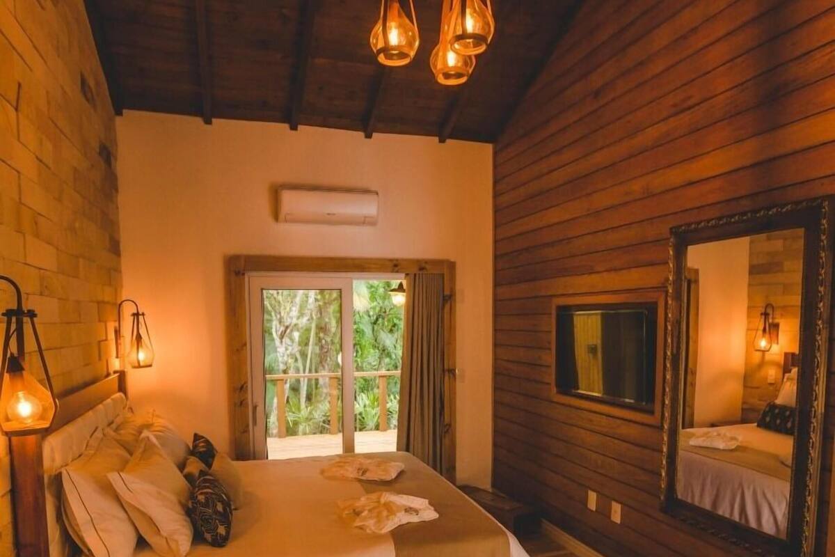 A bedroom with a bed, a wooden floor, a window with curtains, a mirror, and a ceiling light fixture.