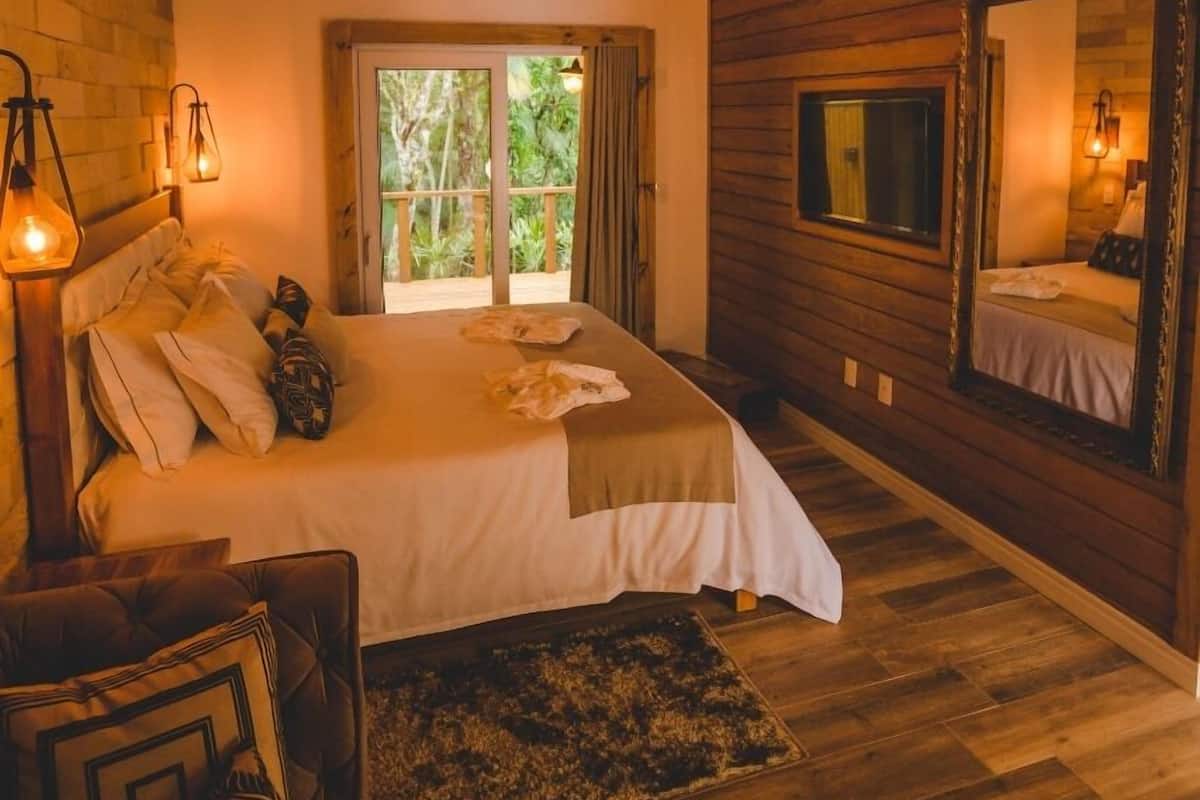 A cozy bedroom with a wooden ceiling, a large bed, a window with curtains, a mirror, and a small table with a lamp.