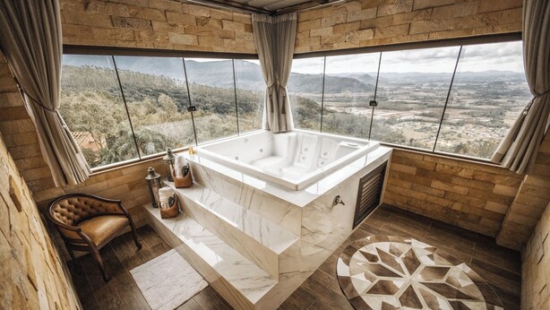 Une salle de bain dotée d’une grande baignoire, d’une chaise en bois, d’une petite table avec des articles de toilette et offrant une vue sur la campagne grâce à de grandes fenêtres.