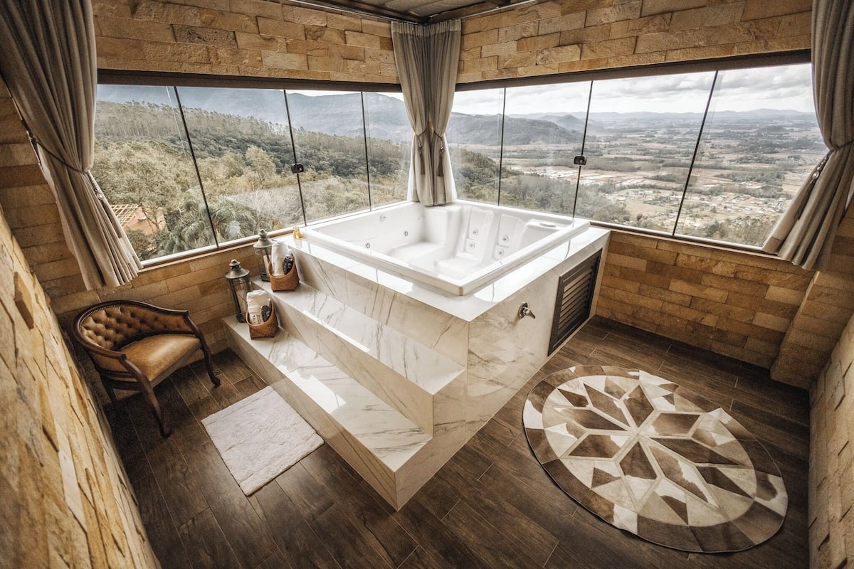 A bathroom with a large bathtub, a wooden chair, a small table with toiletries, and a view of the countryside through large windows.