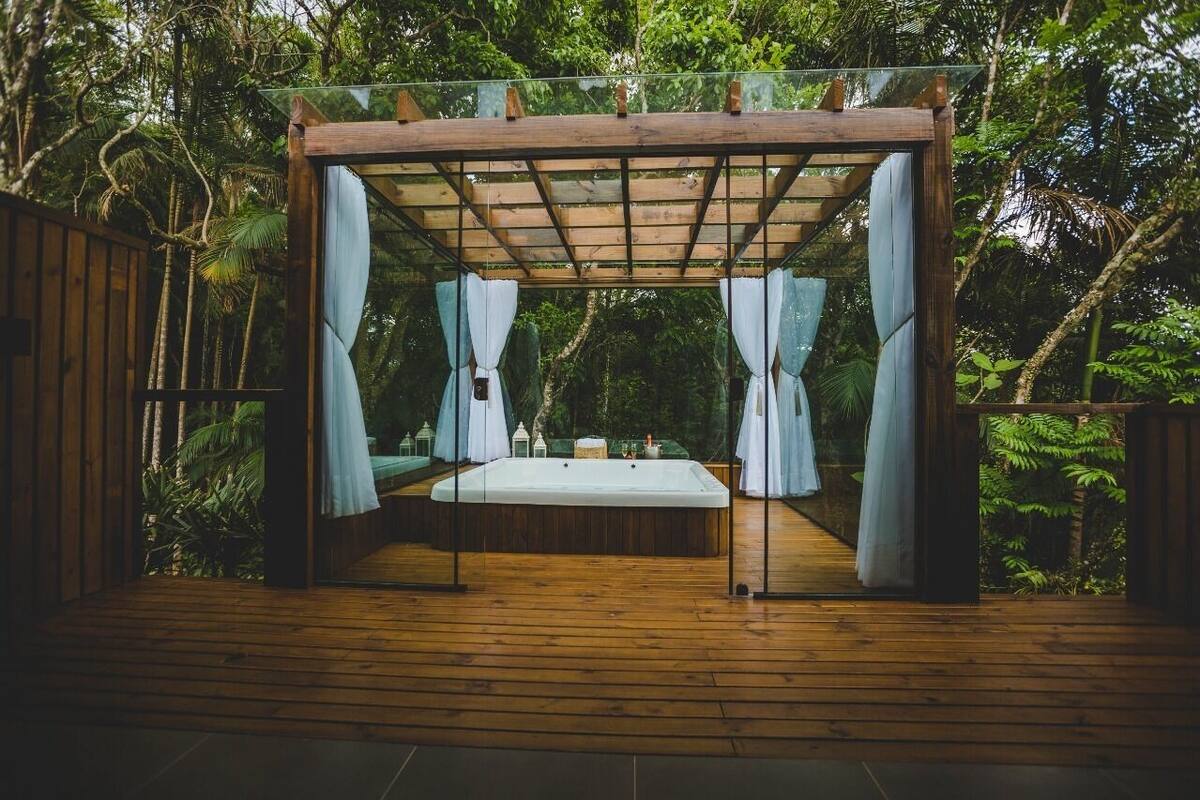 A wooden deck with a glass enclosure, a bed, and curtains.