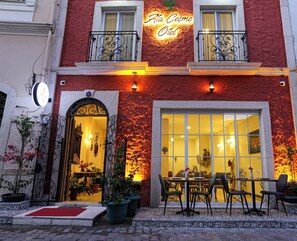 L’entrée d’un hôtel avec une façade rouge, une grande fenêtre en verre, des places assises à l’extérieur et une enseigne indiquant « Atto Cesme Otel ».