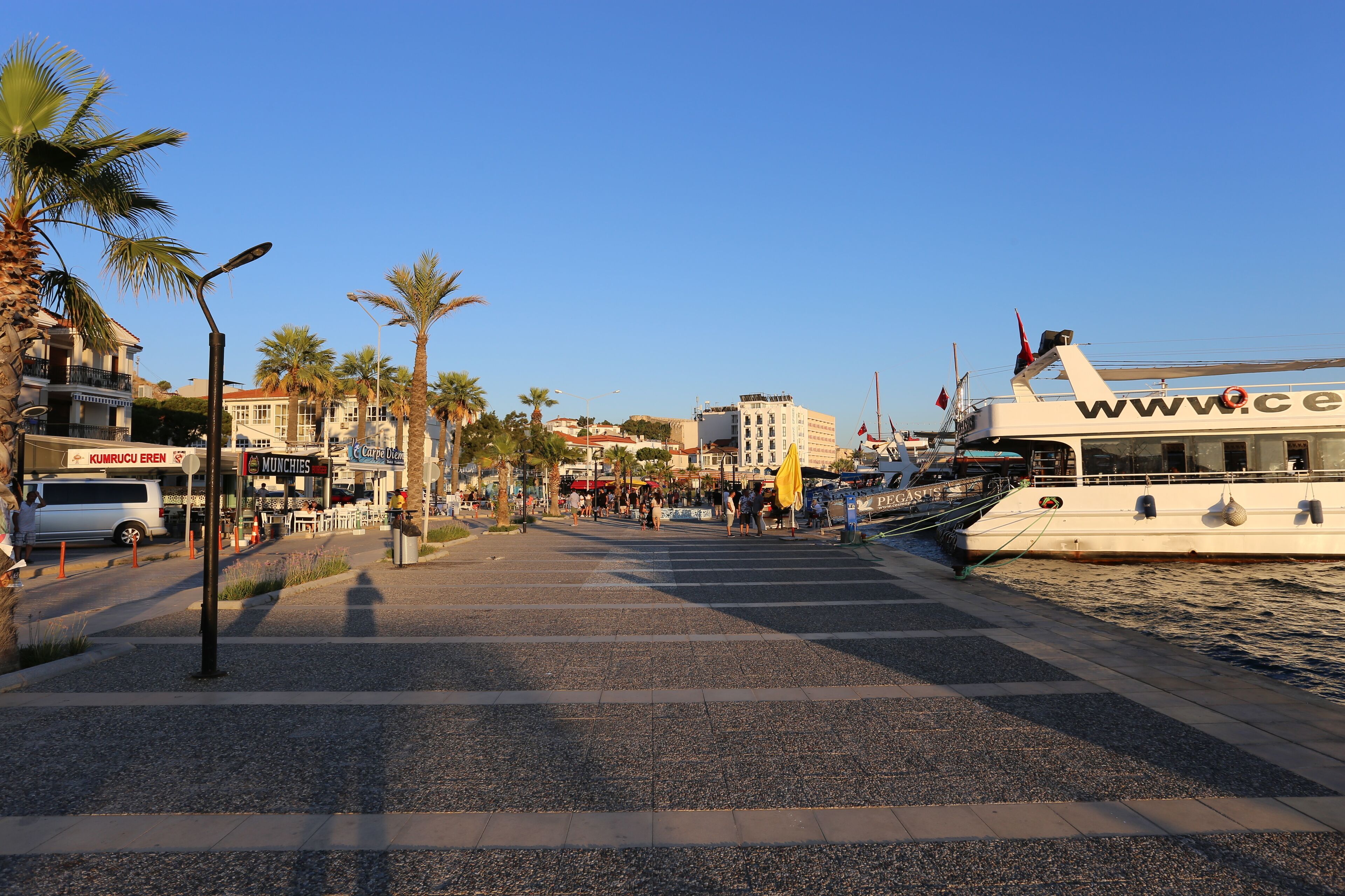 A waterfront promenade with a boat docked on the right, palm trees lining the path, and various shops and buildings in the background.