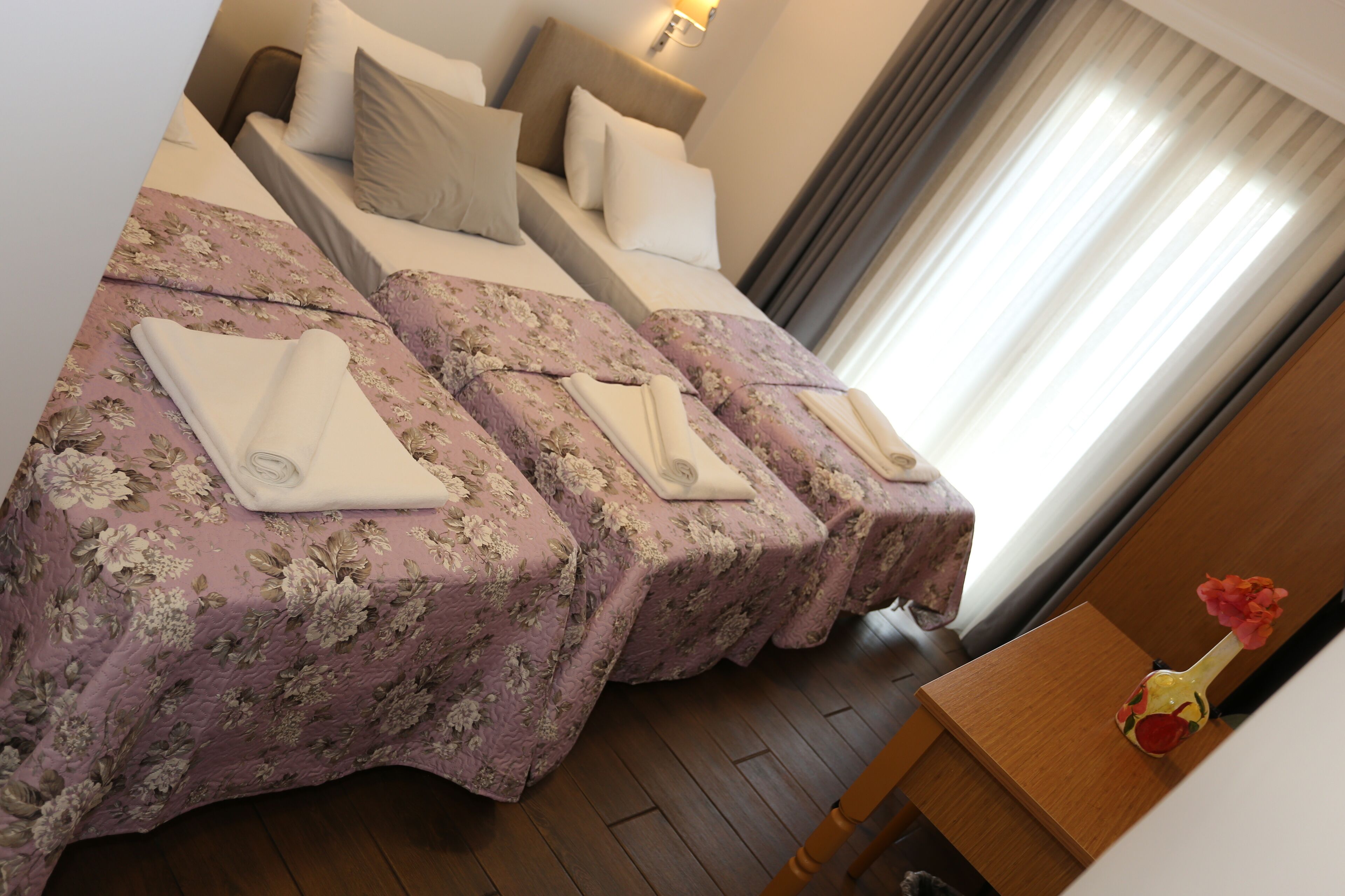 A hotel room with two beds, each with a floral bedspread and white towels. There is a wooden nightstand with a vase of flowers.