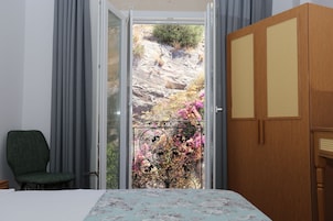 Un dormitorio con cama, una silla, un armario de madera y un balcón con vistas a terreno rocoso y plantas en flor.
