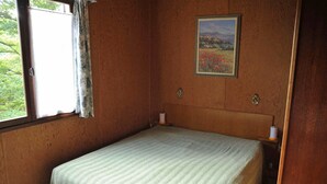 Une petite chambre avec un lit simple, une tête de lit en bois, un tableau représentant un paysage encadré au mur et une fenêtre avec des rideaux.
