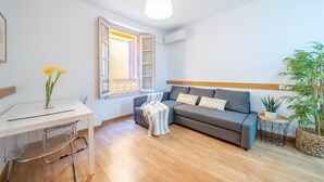 Un salon moderne avec un canapé gris, une petite table d’appoint, un bureau transparent avec une chaise, une fenêtre dont les volets sont ouverts et un luminaire suspendu.