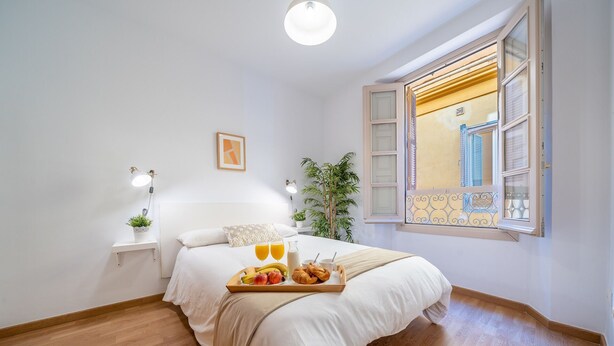 Une chambre à coucher avec un lit, une table de chevet, une lampe, une fenêtre et un plateau contenant des fruits et des pâtisseries.