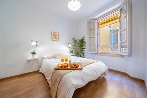 A bedroom with a bed, bedside table, lamp, window, and a tray with fruits and pastries. - Sunset Malaga Apartments - Mitjana 3.2 (Málaga)