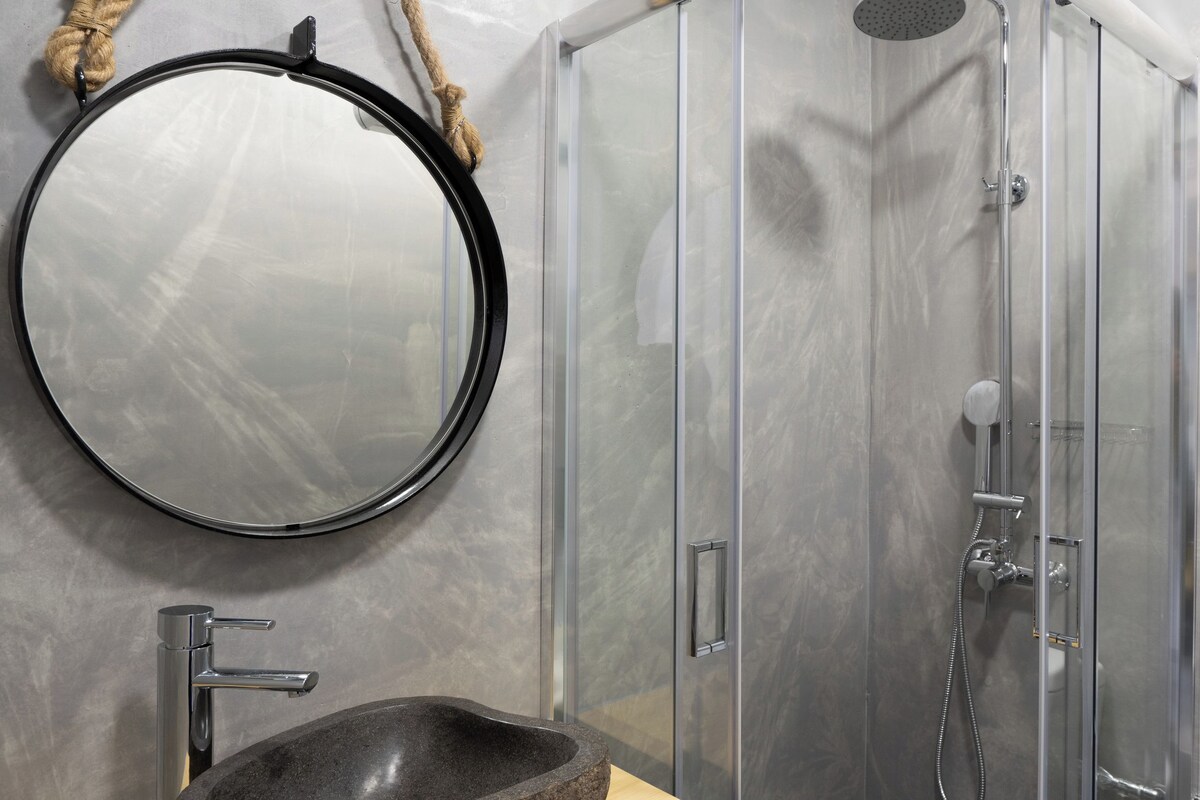 A bathroom with a stone basin, a round mirror, and a glass-enclosed shower.