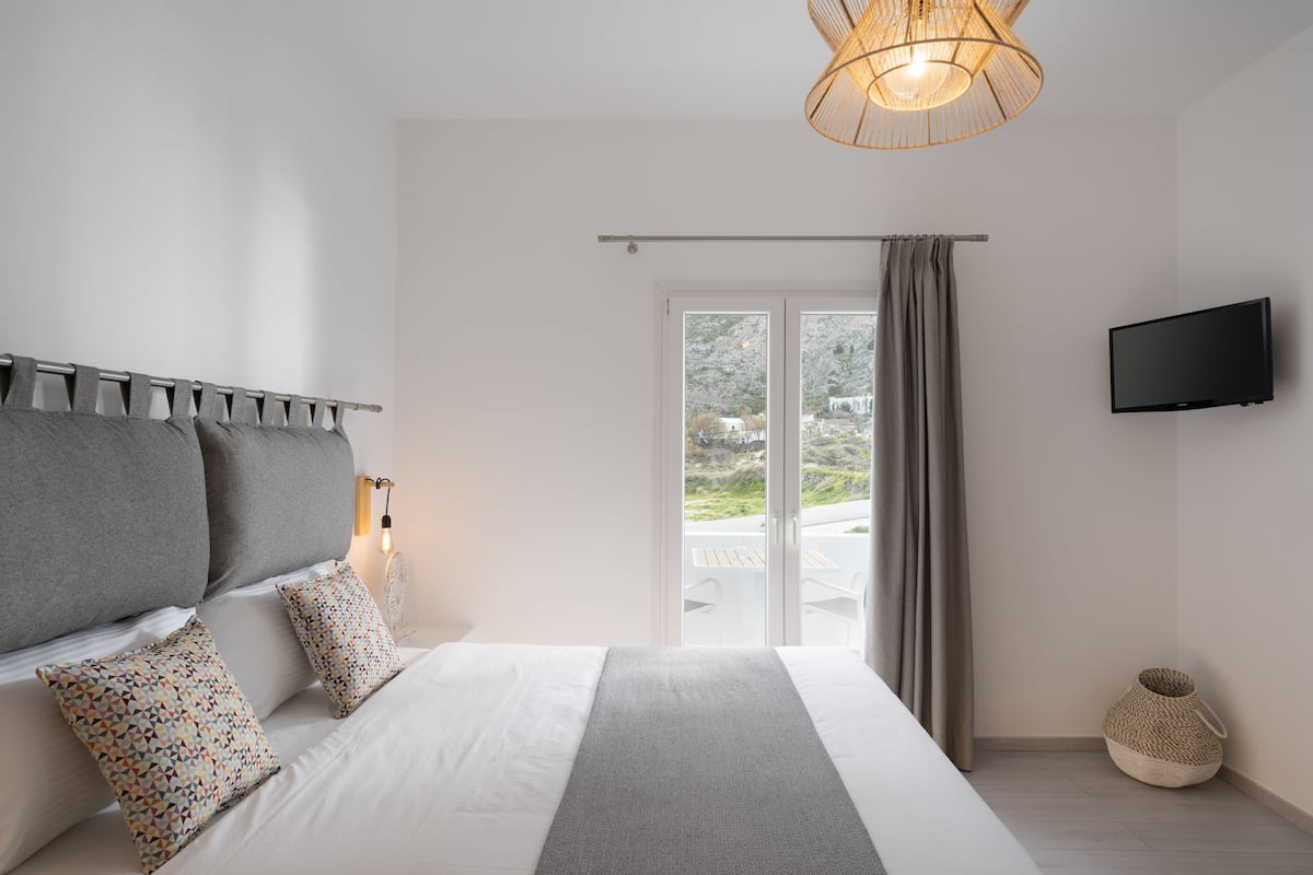 A bedroom with a bed, pillows, a bedside table, a TV, and a view of the outdoors.