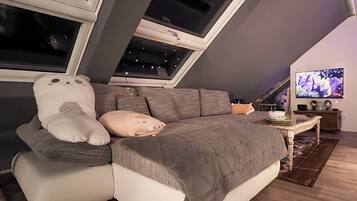 Ein gemütliches Schlafzimmer im Loft-Stil mit schrägem Dach, einem großen Fenster, einem Bett mit grauer Decke und einem weichen weißen Kissen sowie einem Fernseher auf einem Schrank.