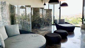 Un área de descanso moderna con un sofá grande y redondo, además de dos opciones de asientos más pequeñas y redondeadas, una pared de mármol y vistas de la ciudad a través de amplias ventanas.
