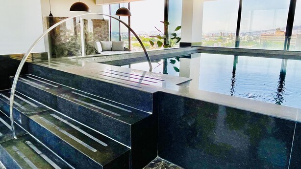 Una piscina interior moderna con barrera de cristal, una escalera curva y vistas a la ciudad.