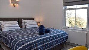 Una cama bien tendida con ropa de cama a rayas, una lámpara de pared y una ventana con vistas a árboles.