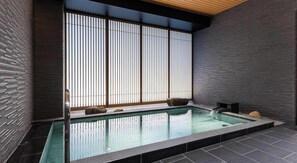 A modern indoor pool with a minimalist design, featuring a large window with vertical blinds, a stone bench, and a small hot tub area.