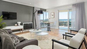 A modern living room with a grey sofa, a white coffee table, and a TV on a white stand. There is a balcony with a glass door and a view of the city.