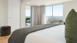 A modern hotel room with a large bed, a view of the sea, and a balcony with a table and chairs.