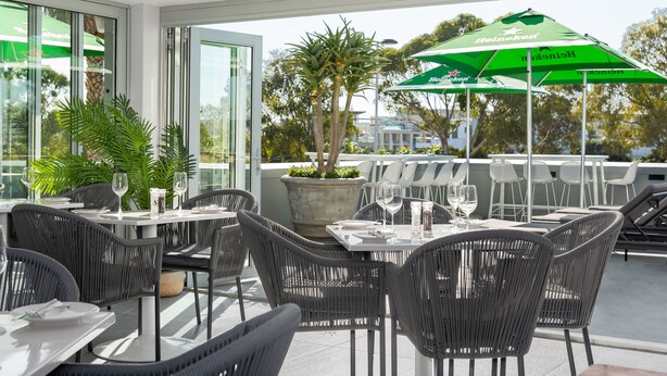 An outdoor dining area with wicker chairs and tables, a large green Heineken umbrella, and a view of the cityscape.