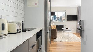 A modern kitchen with a white countertop, a built-in oven, and a refrigerator. There is a dining area with a small table and a chair, and a large window offering a city view. - Point Break | Sleek Studio | Mountain Views (Cape Town)