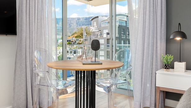 A modern room with a wooden table, two clear chairs, a black lamp, and a view of buildings through large windows. - Point Break | Sleek Studio | Mountain Views (Cape Town)