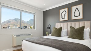A bedroom with a large window, a bed with white linens, green pillows, and a grey throw blanket. - Point Break | Serviced Apartment | Pool Access (Cape Town)