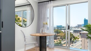 A modern room with a round table, clear chairs, a vase with flowers, a large mirror, and a city view through large windows. - Point Break | Serviced Apartment | Pool Access (Cape Town)