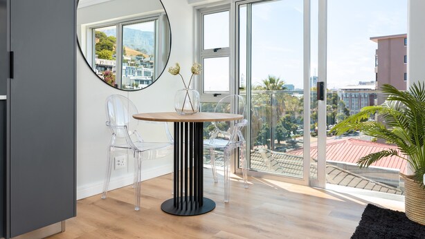 Una habitación moderna con una mesa redonda, sillas traslúcidas, un espejo grande y vistas a la ciudad a través de amplias ventanas.