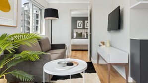 Una habitación de hotel moderna con un sofá gris, una pequeña mesa blanca, una lámpara de pie negra, un televisor de pantalla plana y un ventanal con vistas a un edificio.