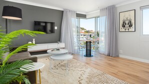 Una sala moderna con un sofá gris, una mesa de centro blanca, una mesita redonda pequeña, un televisor montado en la pared y un balcón con vistas a edificios.