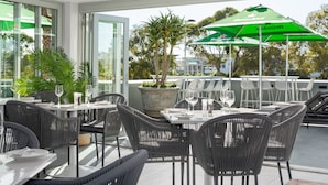 An outdoor dining area with wicker chairs and tables, a large green Heineken umbrella, and a view of the cityscape. - Point Break | Ocean Views | Walk to VandA | Pool (Cape Town)