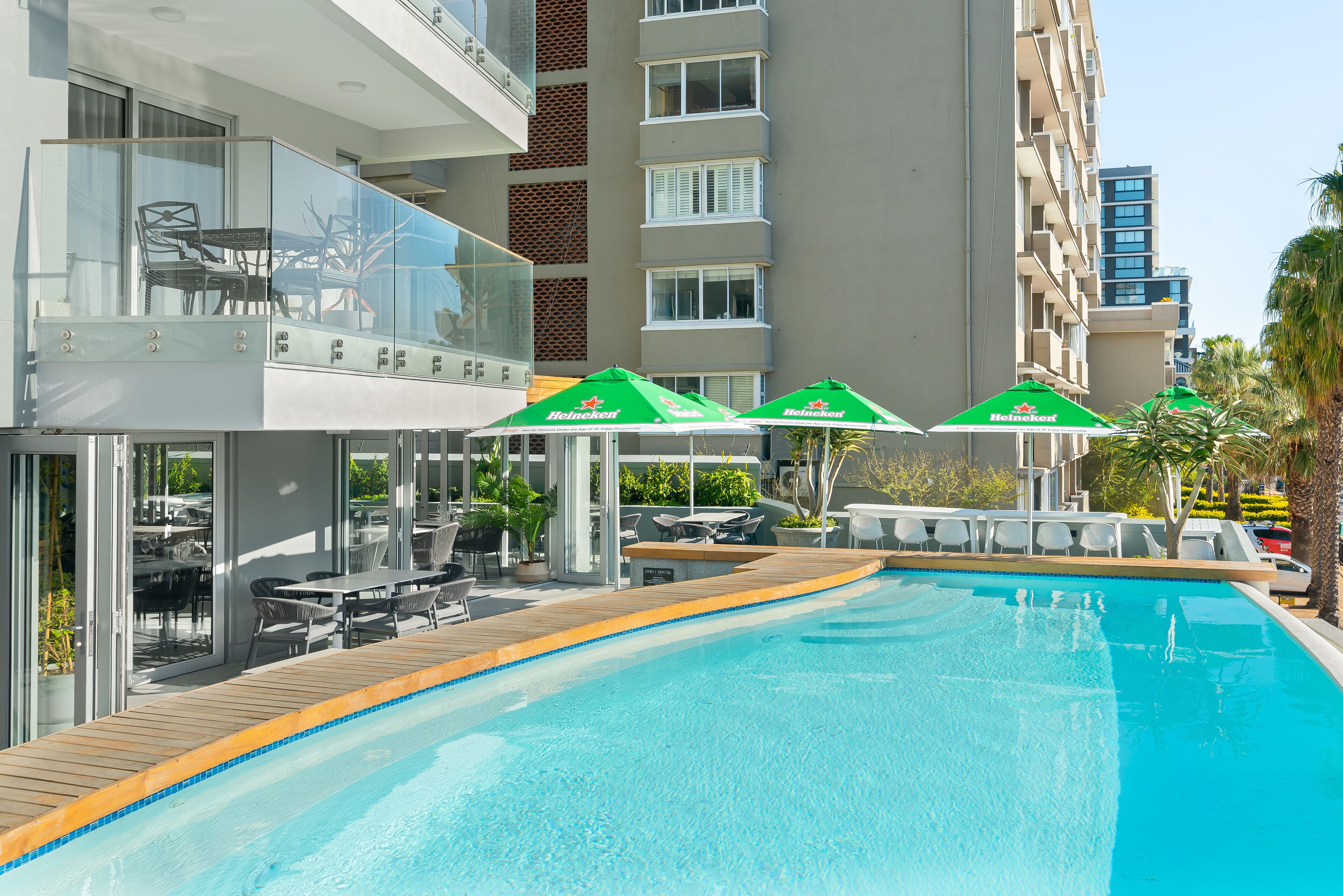 A swimming pool with a wooden deck, outdoor seating, and Heineken umbrellas.