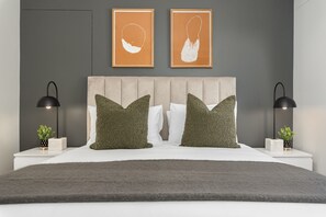 A modern bedroom with a tufted headboard, two green pillows, a white bedspread, and two bedside tables with black lamps and small potted plants.