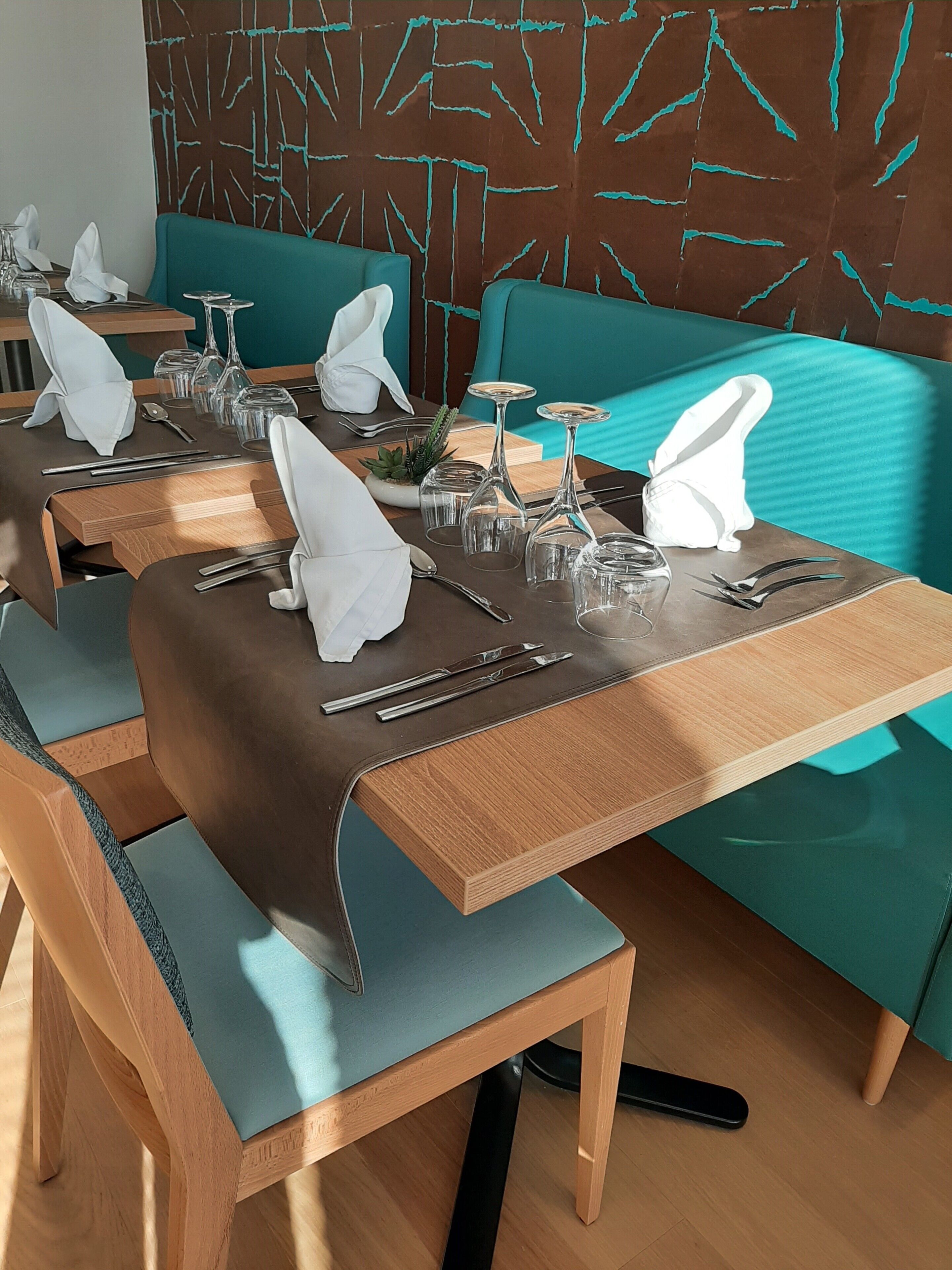 Un espace repas avec des banquettes recouvertes d’un tissu turquoise, des tables en bois et de la verrerie transparente.