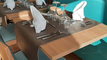 Un espace repas avec des banquettes recouvertes d’un tissu turquoise, des tables en bois et de la verrerie transparente.