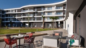Un'area salotto esterna moderna con un divano bianco e poltrone rosse, un tavolo con teiera e vista su un edificio a più piani con balconi.