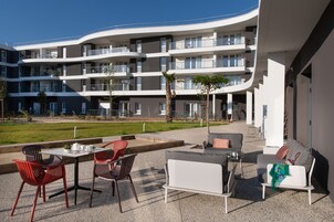 Un área de estar moderna al aire libre con un sofá blanco y sillas rojas, una mesa con tetera, y vistas a un edificio de varios pisos con balcones.