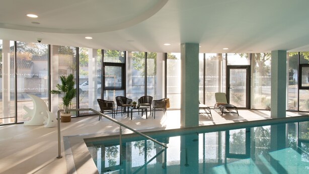 Una piscina interna con pareti in vetro trasparente, un’area relax con sedie e un’ampia pianta in vaso.