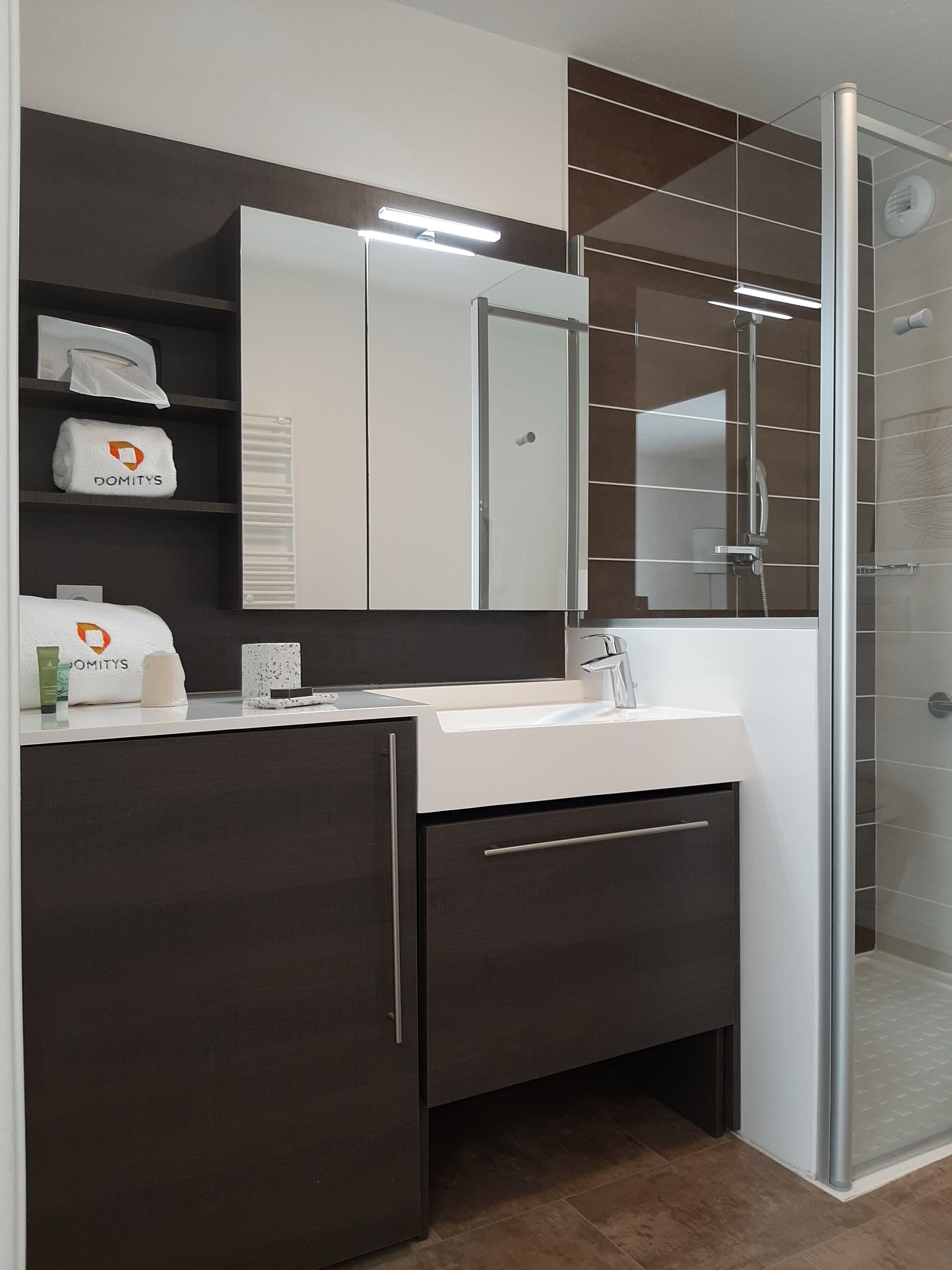 Un bagno moderno con un lavandino bianco, mobili scuri, uno specchio grande e un box doccia in vetro.