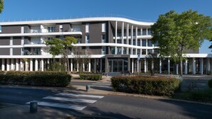 Un edificio moderno a più piani con facciata curva, con l'insegna "BASALTIK". Presenta balconi, un ingresso centrale ed è circondato da vegetazione.