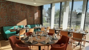 Zona pranzo moderna con tavoli rotondi apparecchiati per quattro persone, circondati da sedie rosse. In secondo piano si intravede una divisa turchese, e la stanza presenta ampie finestre con tende.