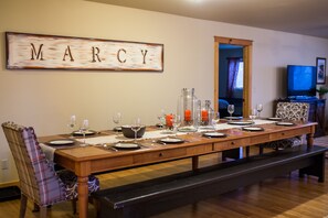 A dining room with a long wooden table set for a meal, a plaid chair, and a bench. A sign with the name "MARCY" is on the wall.