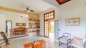 A spacious living area with a dining table, wooden chairs, and a sofa. There are ceiling fans, a large window, and a door leading outside.
