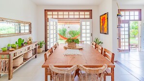 A spacious dining area with a wooden table and wicker chairs, a large mirror, and a view of a garden through open doors.
