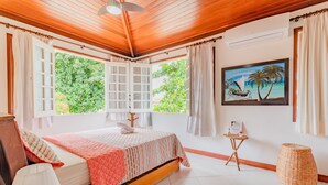 A bedroom with a wooden ceiling, a bed with a pink quilt, a painting of a beach scene, and a wicker stool.