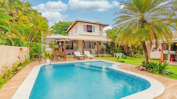 A rectangular swimming pool with a tiled deck, surrounded by a well-maintained garden and a two-story house with a tiled roof.