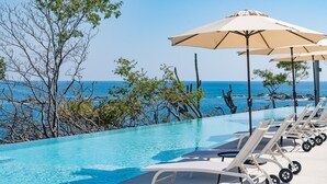 A poolside area with lounge chairs and umbrellas, overlooking a clear blue sea with trees and cacti in the background. - Ocean View Penthouse with Rooftop Terrace (Huatulco)