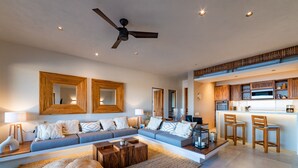 A modern living room with a sofa, coffee table, and ottomans. There is a kitchen area with a bar and stools. - Ocean View Penthouse with Rooftop Terrace (Huatulco)