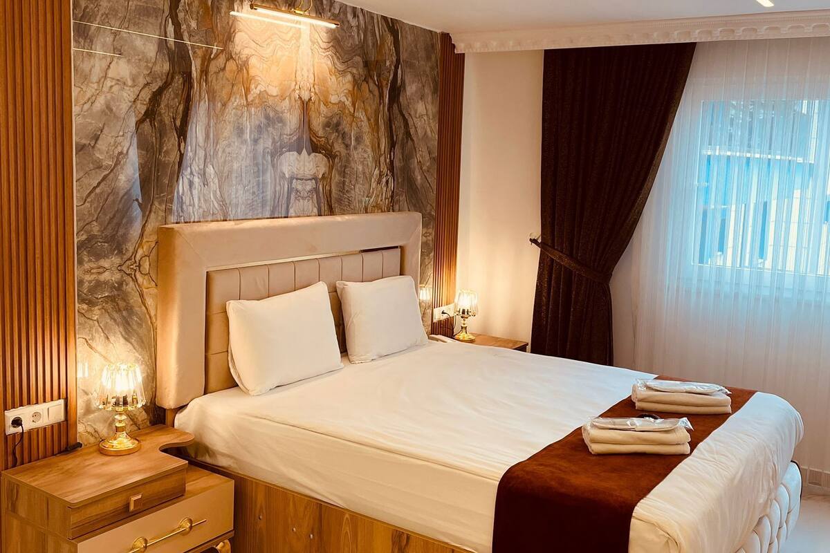 A modern bedroom with a marble-patterned wall, a wooden nightstand with a drawer, a bedside table with a lamp, and a bed with a brown throw blanket.